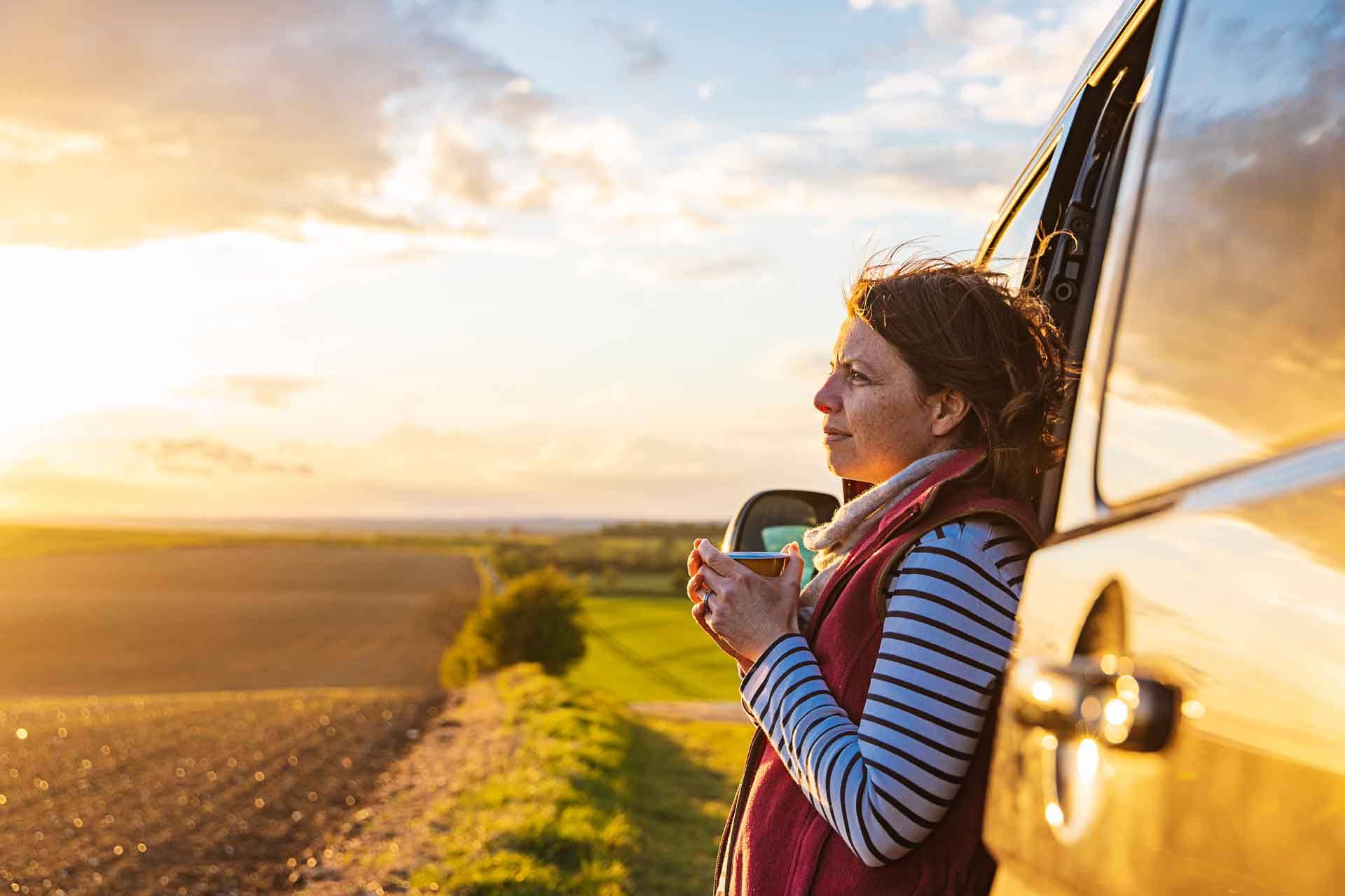 A woman leaning on a truck watching the sunset over a farm.